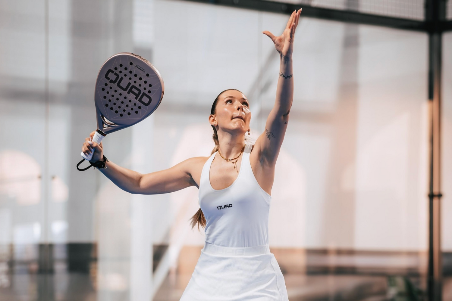 Woman playing paddle tennis in a modern indoor setting, holding a paddle with 'Quad' branding.